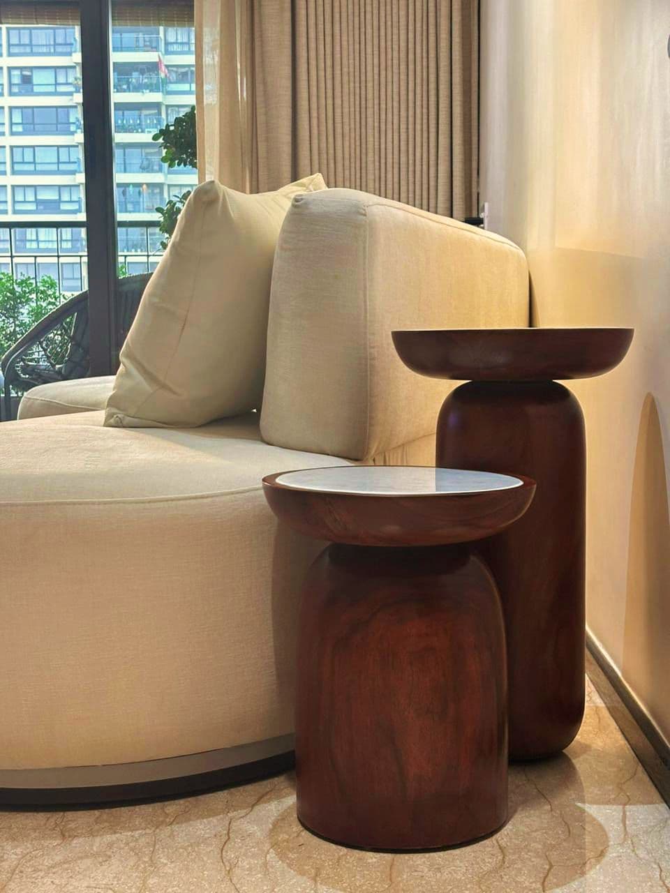 Two sculptural pedestal wood side tables beside a cream sofa, with a city view through the window.