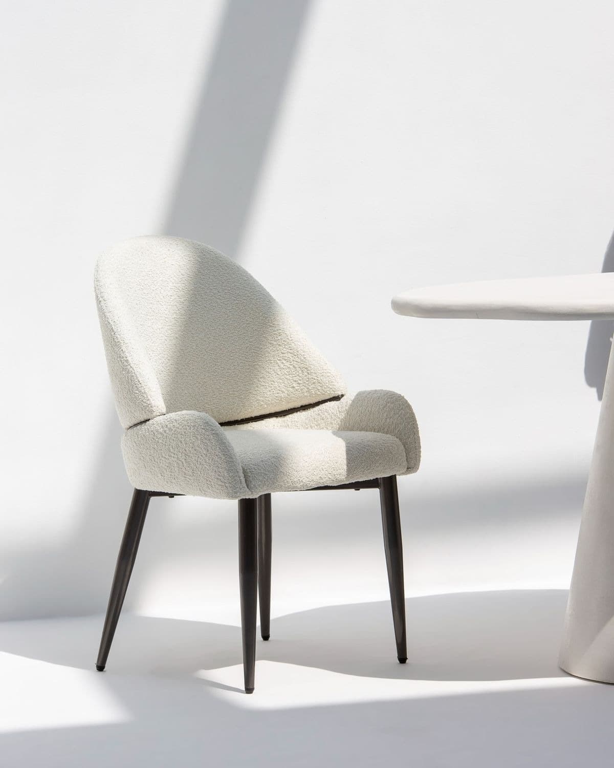 ÉLÉA premium dining armchair with curved neutral upholstery and slim matte-black frame in a bright modern interior.