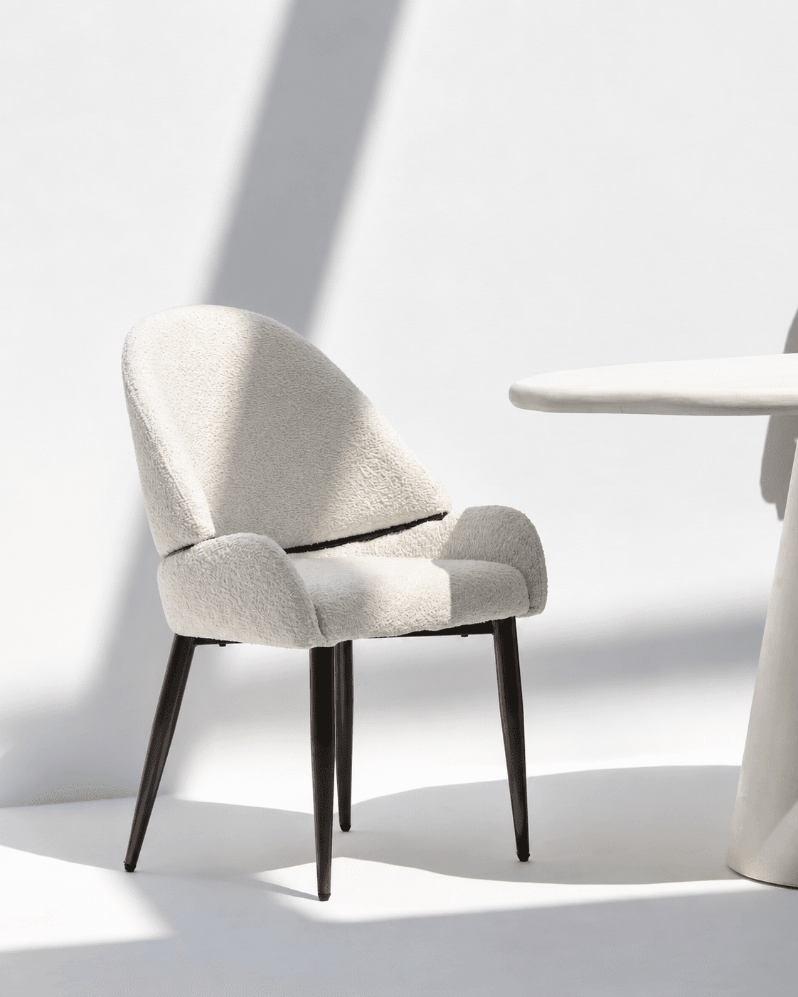 ÉLÉA premium dining armchair with curved neutral upholstery and slim matte-black frame in a bright modern interior.